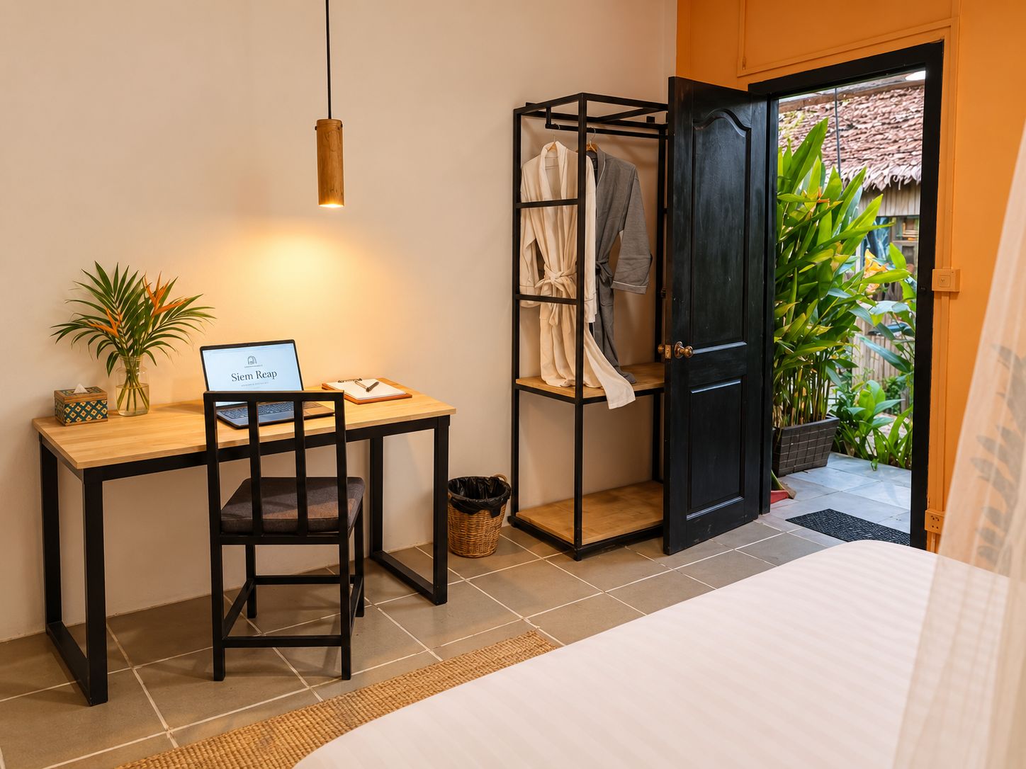 Quarto com cama, escrivaninha com laptop, cadeira, mesinha com planta, guarda-roupa preto e porta que dá para um jardim.