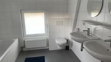 A modern bathroom with a bathtub, two sinks, a toilet, and a window.