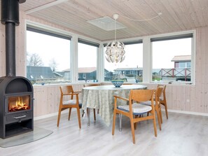 A room with a wood-burning stove, a round dining table with chairs, large windows, and a hanging light fixture. - 7 Person Holiday Home in Juelsminde (Horsens)