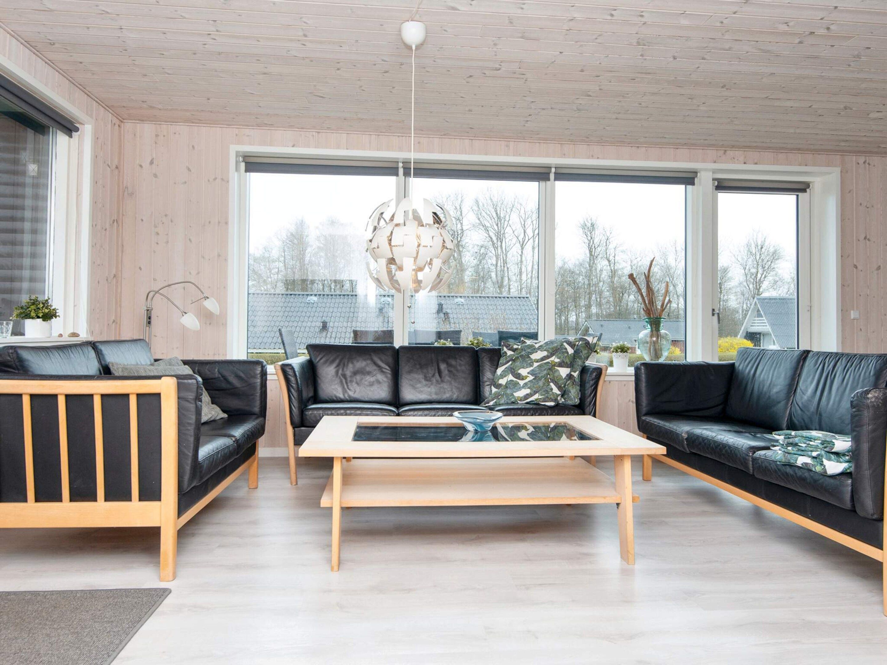 A modern living room with a wooden ceiling, large windows, a central coffee table, and two black leather sofas.