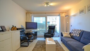 Ett vardagsrum med en soffa, en tv, ett skrivbord och en stol. Det finns ett fönster med gardiner och utsikt över byggnader utanför.