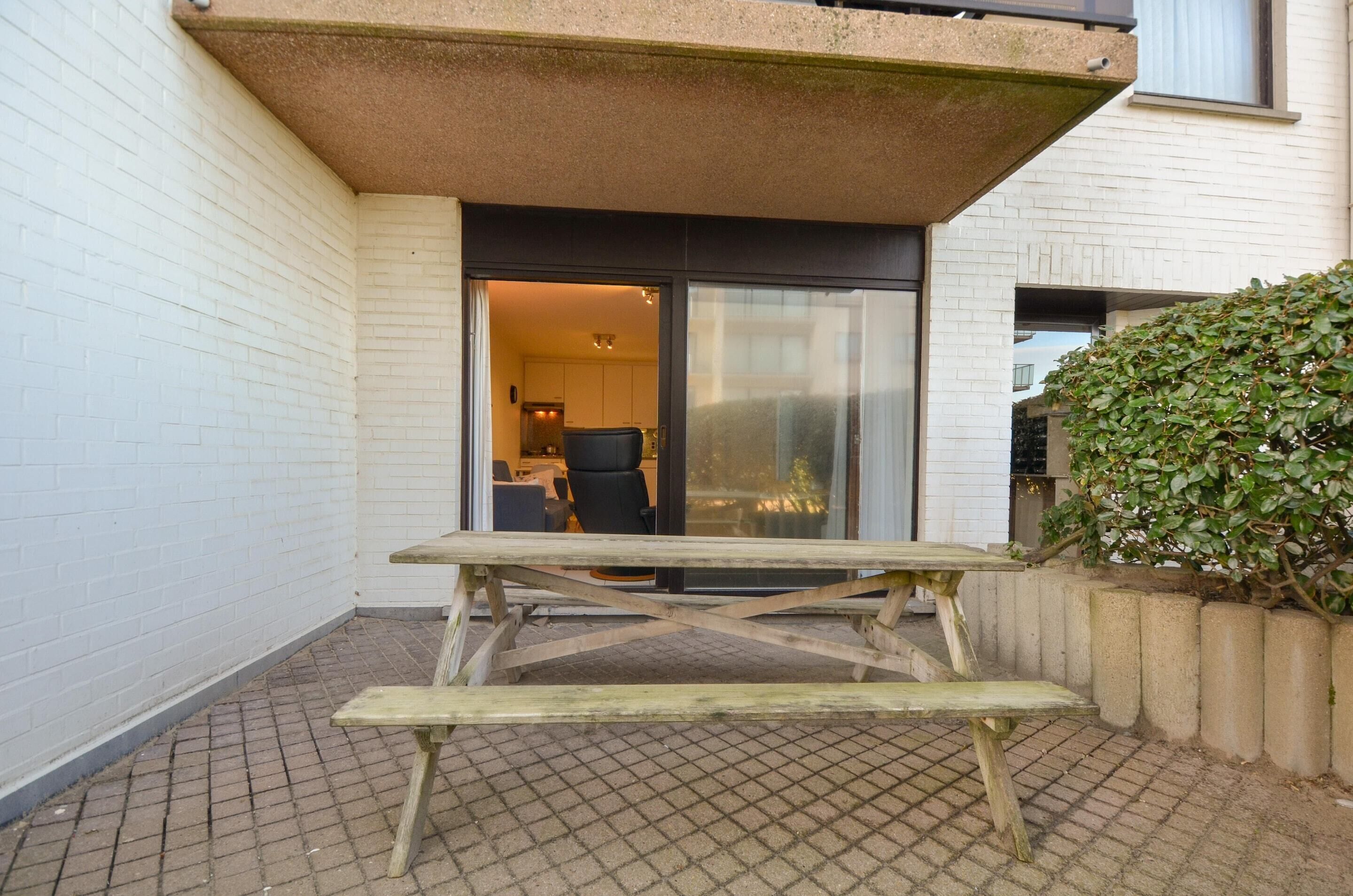A wooden picnic table in a courtyard with a white brick wall and a glass door leading to a room.