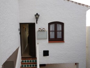 A white building with a dark brown door, a small window with curtains, and a wall-mounted light fixture. - La Perla - South Facing, Sun Morning to Evening (Nerja)