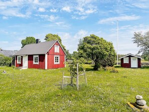 A red wooden house with a grey roof, surrounded by a grassy field with a large tree and a small wooden structure.