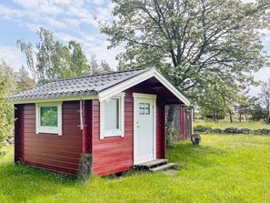 A small, single-story cabin with a red exterior and white trim, situated on a grassy area with trees in the background.