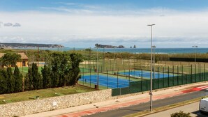 Cancha de tenis con muro de piedra y árboles en primer plano, una carretera con una línea roja, y vista al mar y accidentes geográficos distantes.