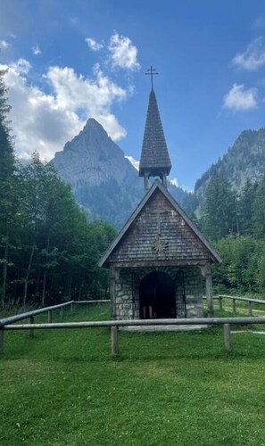 A small stone chapel with a cross on top, surrounded by lush greenery and mountains in the background.
