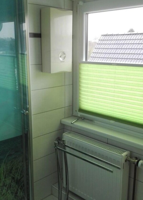 A bathroom with a glass shower enclosure, a window with blinds, a radiator, and a white cabinet.