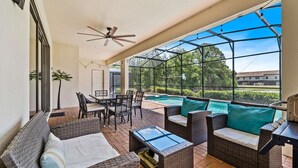 Un patio couvert aménagé avec des meubles en osier, une table en verre et une vue sur une piscine et la verdure environnante.