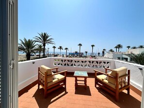 Eine Terrasse mit zwei Holzstühlen und einem kleinen Tisch, mit Blick auf einen Strand mit Palmen und einem klaren Himmel.