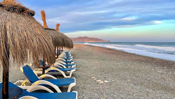 Una playa con sombrillas de paja y sillas reclinables azules mirando al océano.