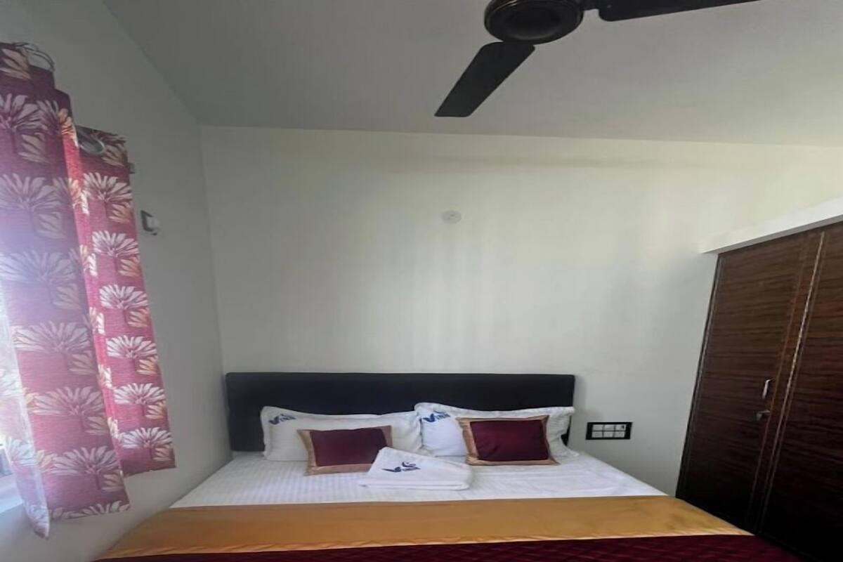 A neatly made bed with a dark headboard, a red and gold bedspread, and a window with red curtains.