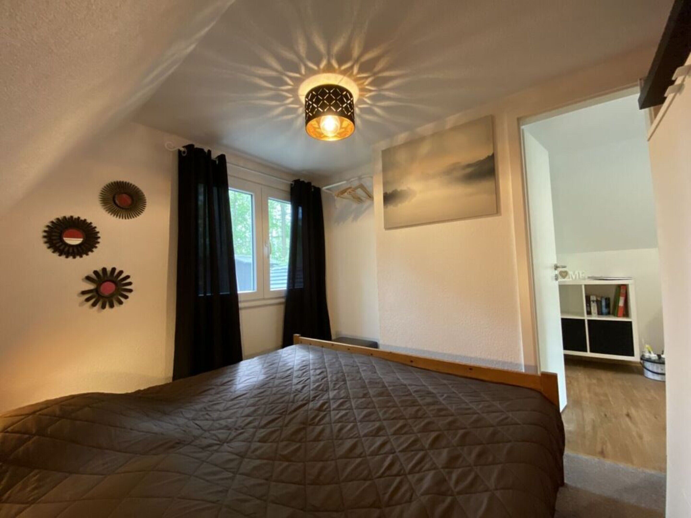 A bedroom with a bed, a window with curtains, a ceiling light fixture, and a painting on the wall.