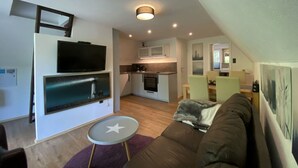 A compact living space with a kitchenette, a flat-screen TV mounted on the wall, a brown sofa, and a small round table. - Holiday Home Seehutte (Malschwitz)