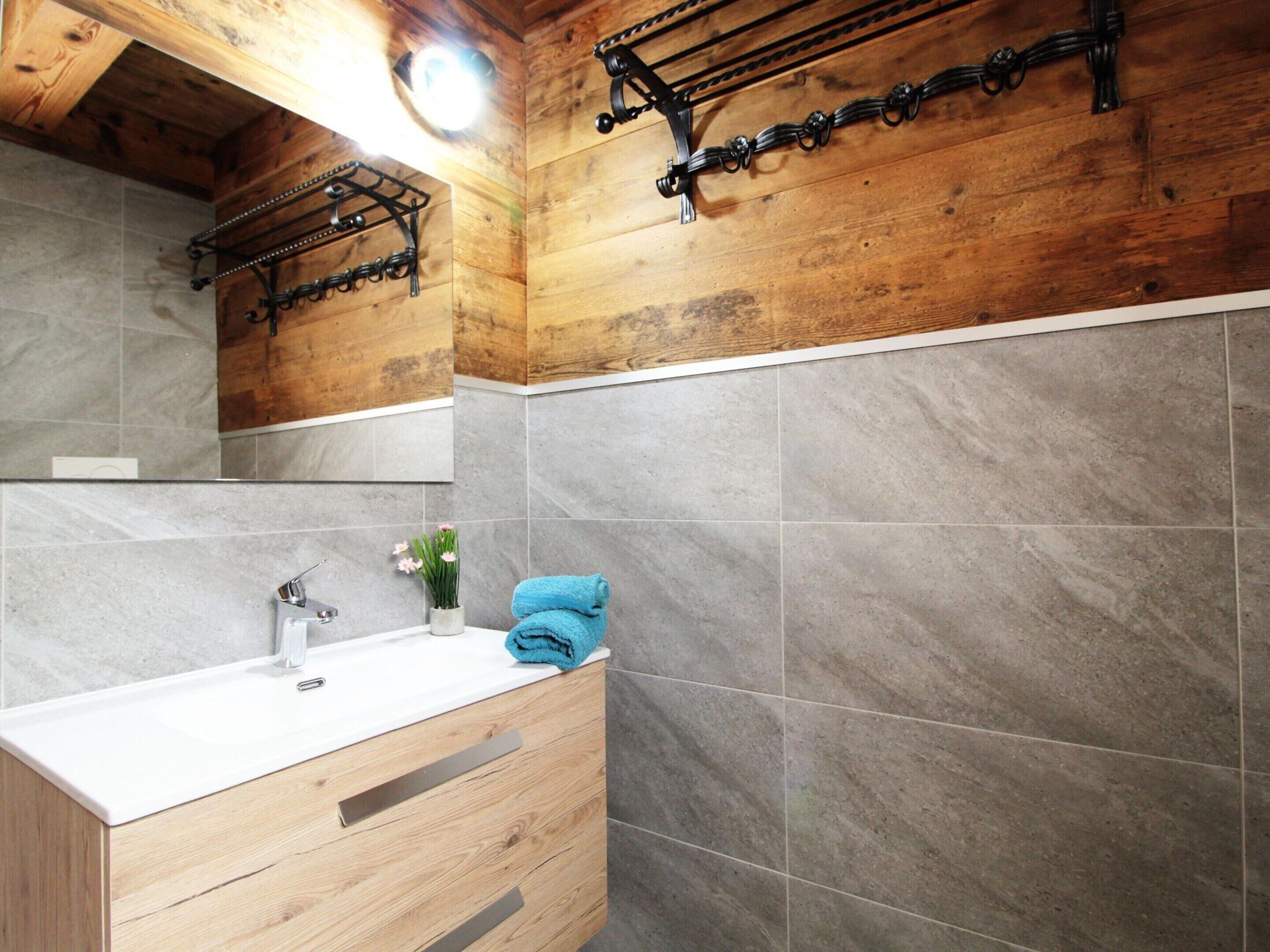 A bathroom with a wooden ceiling, a white sink, grey tiles, a mirror, and a towel rack.