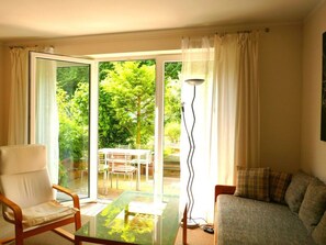 Una sala de estar con puerta de cristal que da a un espacio exterior, una silla blanca, un sofá y una lámpara de pie.