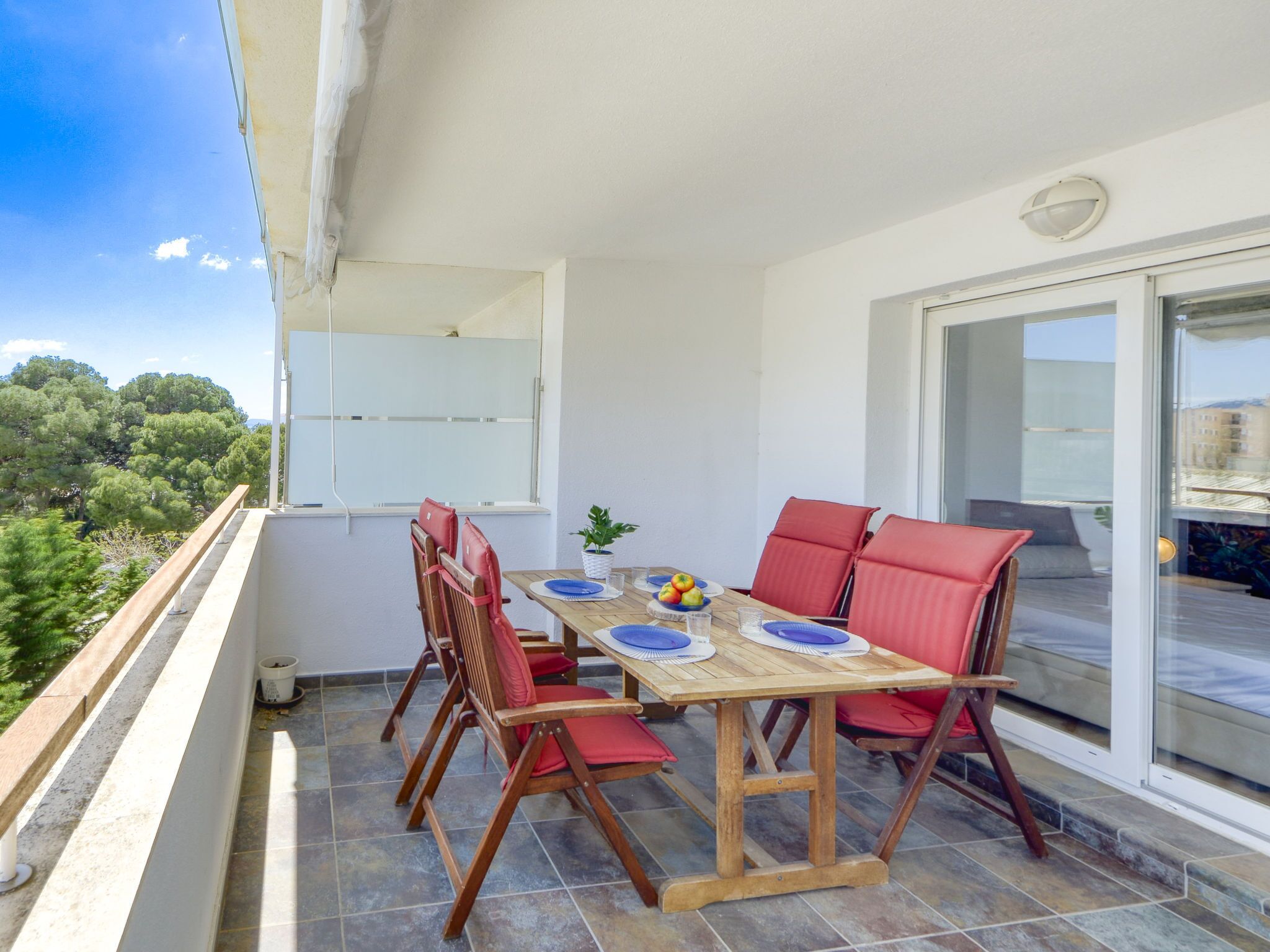 A balcony with a wooden table and red chairs, a potted plant, and a view of trees and buildings.