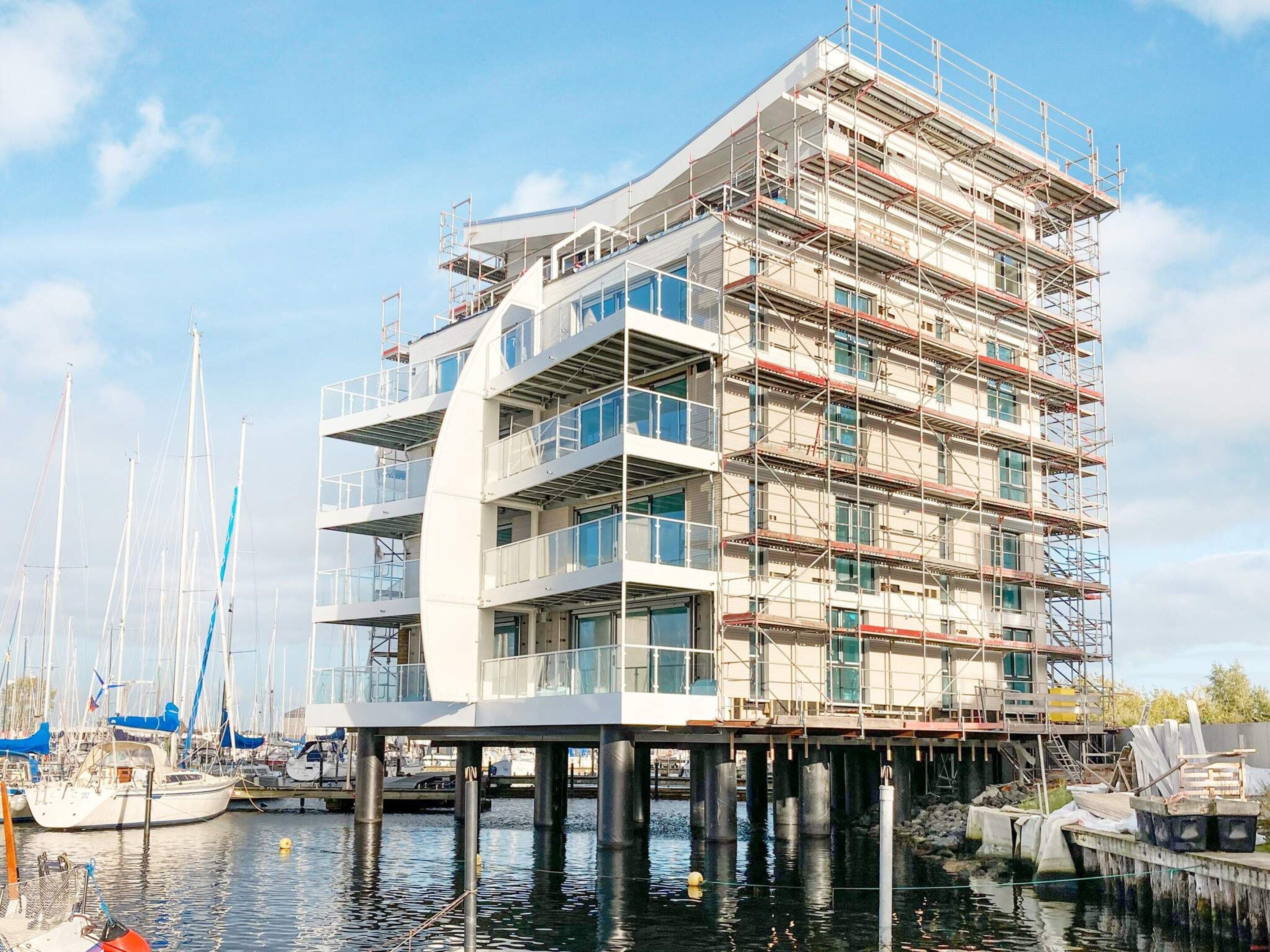 A multi-story building with a modern architectural design, featuring balconies and large windows, situated on stilts over water with boats docked nearby.