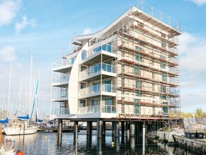 A multi-story building with a modern architectural design, featuring balconies and large windows, situated on stilts over water with boats docked nearby. - 5 Star Holiday Home in Wendtorf (Wendtorf)