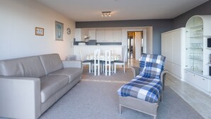 A modern living room with a sofa, a chair, a dining table with chairs, and a kitchen area in the background. - Attractive Studio With a Sea-view (Middelkerke)