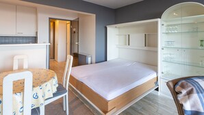 A compact room with a bed, a small table, a chair, and a glass cabinet with shelves. - Attractive Studio With a Sea-view (Middelkerke)