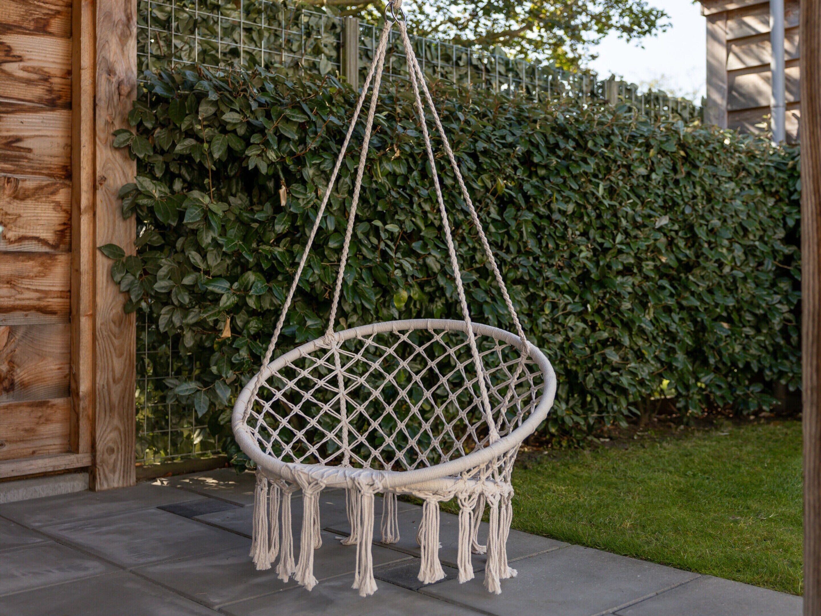 Un fauteuil suspendu en macramé, maintenu par des cordes, avec une clôture en bois et un arbuste en arrière-plan.