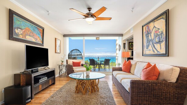 A living room with a television, a wicker sofa, a glass coffee table, and a view of the ocean.