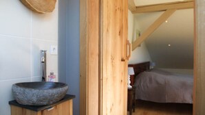 A bathroom with a stone basin, wooden cabinets, and a wooden door leading to a bedroom with a bed and a nightstand. - Villa Carefree Suite (Grassau)