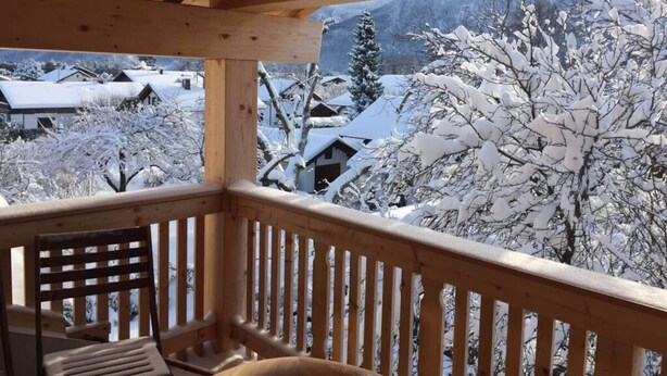 A wooden balcony with a chair, overlooking a snowy landscape with houses and trees. - Villa Carefree Suite (Grassau)