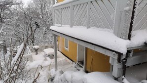 A snow-covered porch with a yellow door, surrounded by snow-laden trees and a white fence.