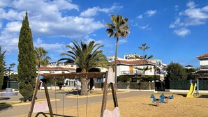 A playground with swings and a slide, surrounded by palm trees and buildings.