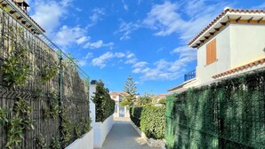 A narrow pathway flanked by a metal fence and a green hedge, leading to a building with a tiled roof and a blue balcony.
