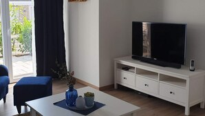 A modern living room with a flat-screen TV on a white TV stand, a blue sofa, a coffee table with decorative items, and a view of the outdoors through a sliding glass door. - Holiday Apartment on the Lindenallee (Bad Rothenfelde)