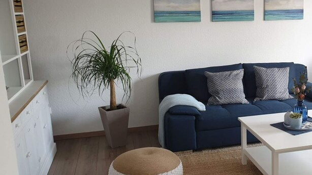 A living room with a blue sofa, a white coffee table, a potted plant, and a small round ottoman. - Holiday Apartment on the Lindenallee (Bad Rothenfelde)
