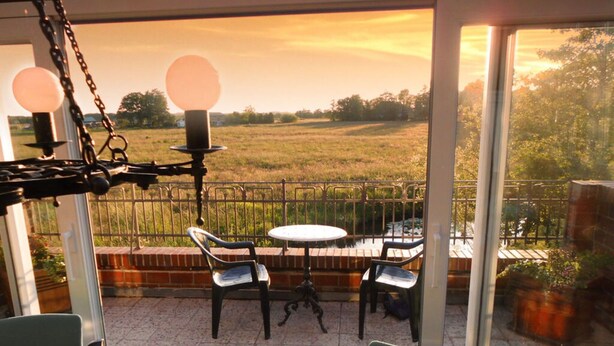 A dining area with a table and chairs, a view of a field through a glass door, and a metal railing. - Pumpe an der Eider (Dellstedt)
