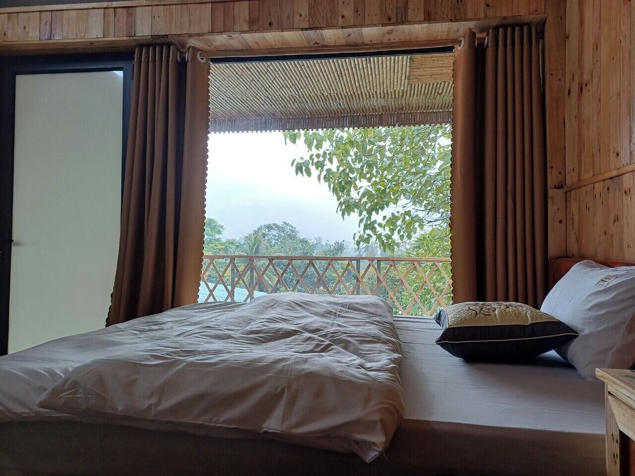 A wooden room with a bed, curtains, and a view of greenery outside.