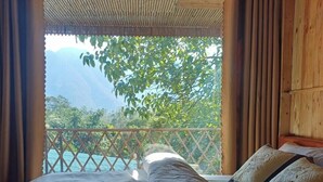 Un lit avec du linge de lit blanc et des coussins décoratifs, une tête de lit en bois et une vue sur un paysage montagneux par une fenêtre ornée de rideaux.