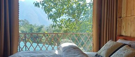 A bed with white linens and decorative pillows, a wooden headboard, and a view of a mountainous landscape through a window with curtains.