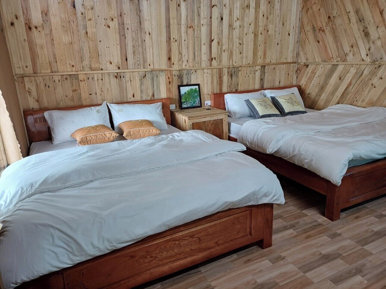 Une chambre avec deux lits en bois, chacun agrémenté de draps blancs et de coussins décoratifs. Une petite table de chevet en bois, sur laquelle est posée une photo encadrée, se trouve entre les deux lits.