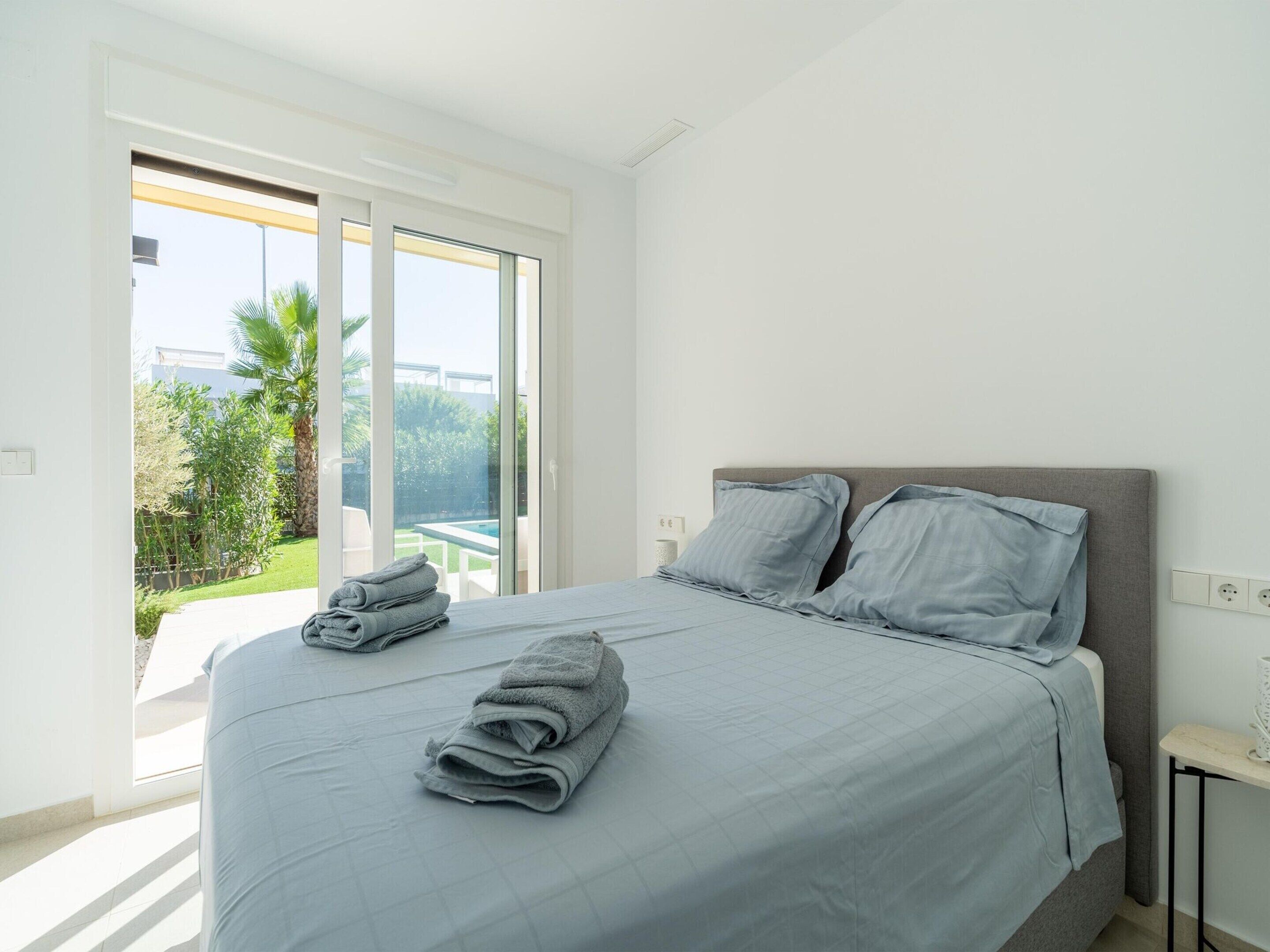 Een slaapkamer met een bed, twee kussens, een nachtkastje en uitzicht op een zwembad en groen buiten.