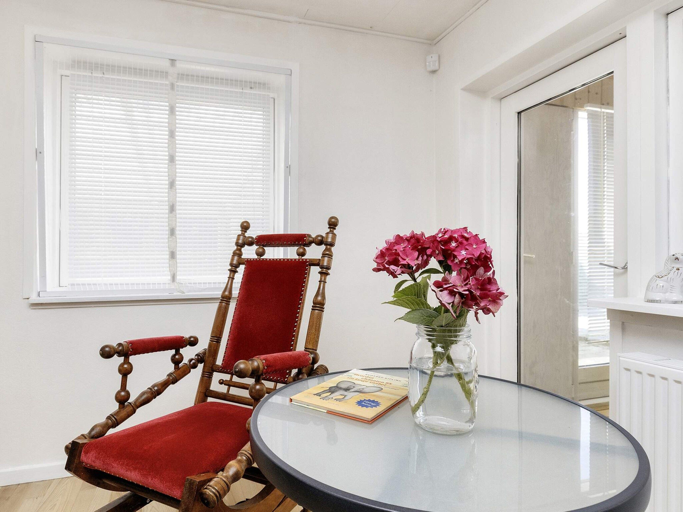 Quarto com cadeira de madeira e estofamento vermelho, mesa de vidro com um livro e um vaso de flores, e janela com persianas.