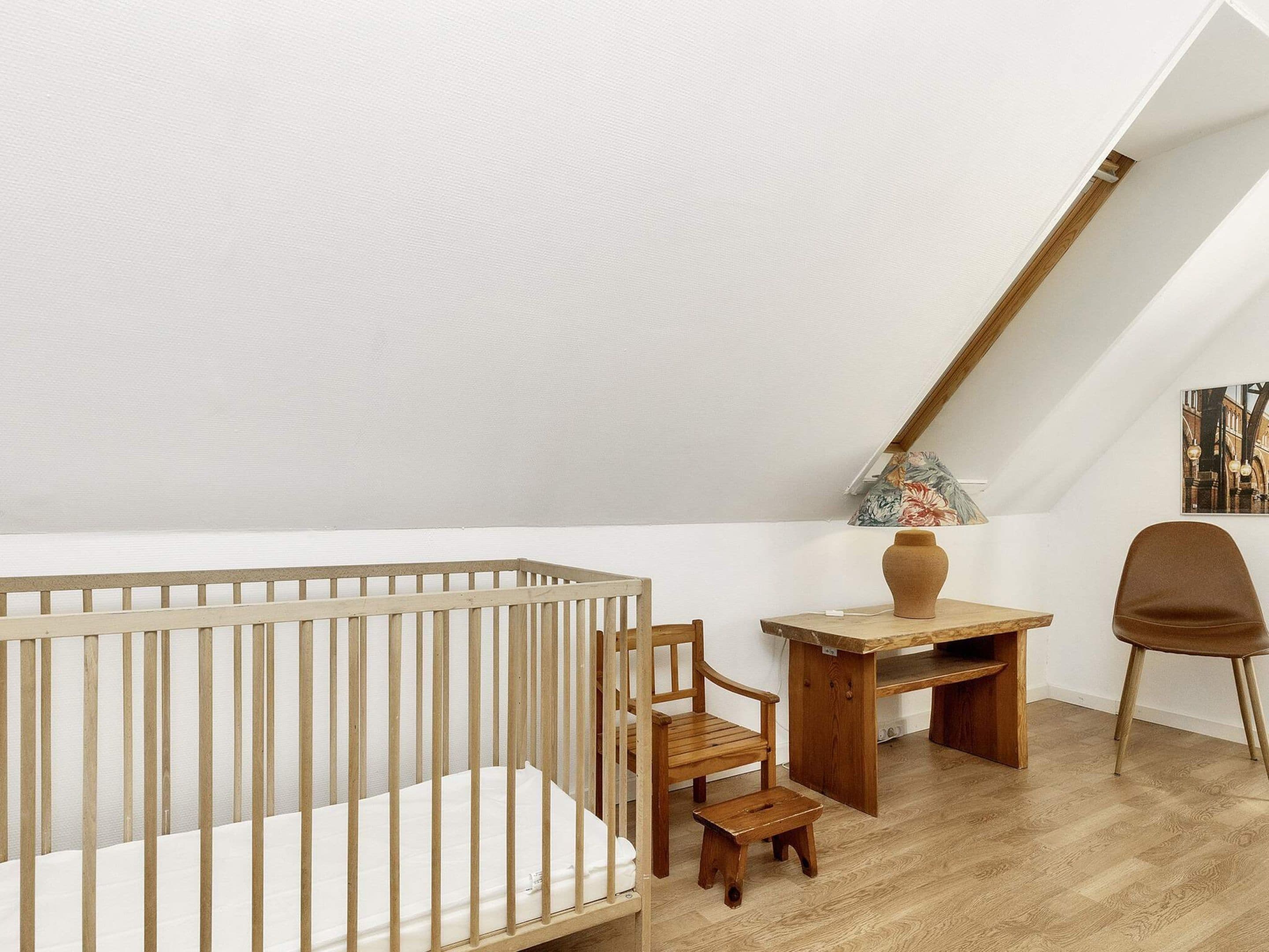 A high-ceilinged room with a wooden crib, a small wooden table, a chair, and a wooden stool.