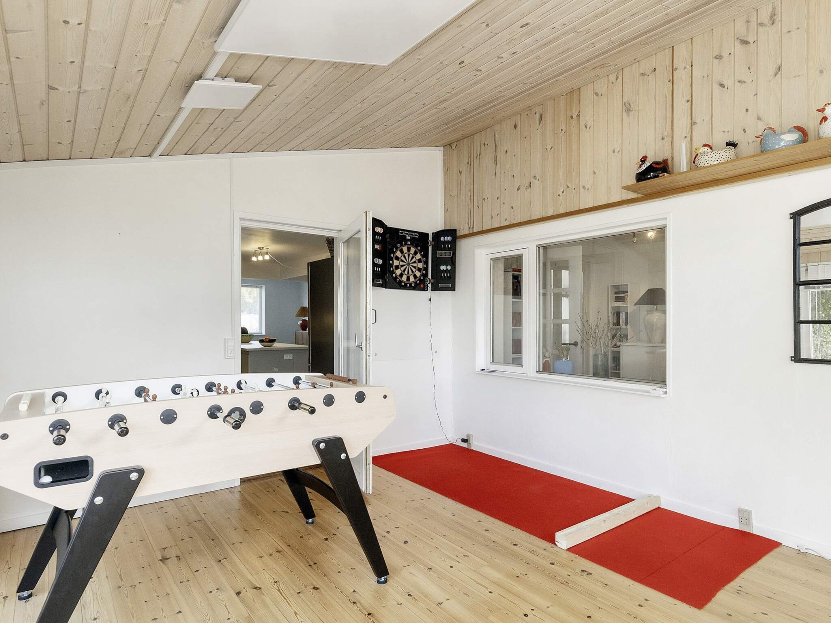 A room with a foosball table, a dartboard, and a window with a view of a kitchen.