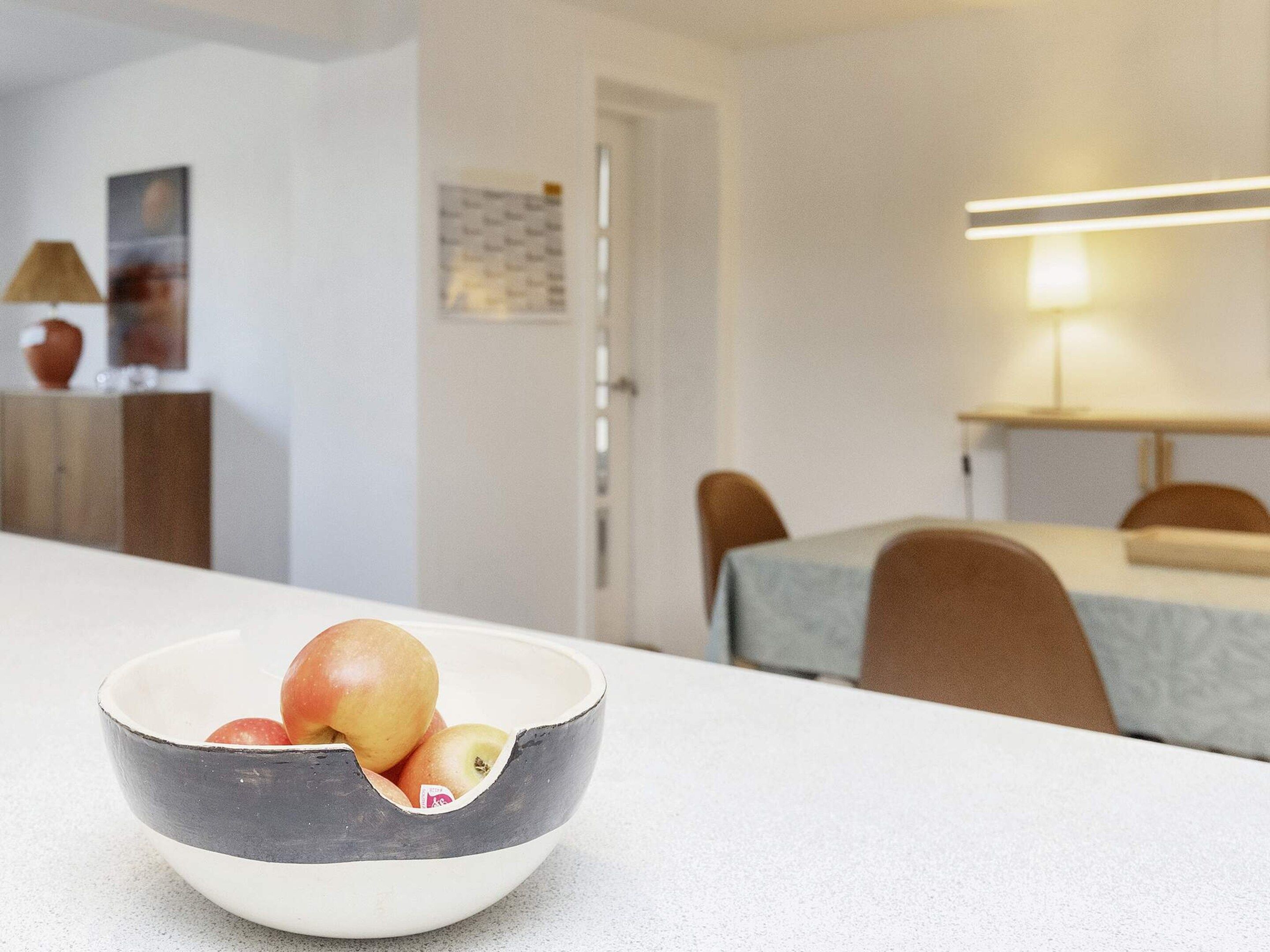A dining area with a table and chairs, a bowl of fruit on the table, and a lamp in the background.