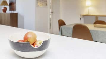 A dining area with a table and chairs, a bowl of fruit on the table, and a lamp in the background.