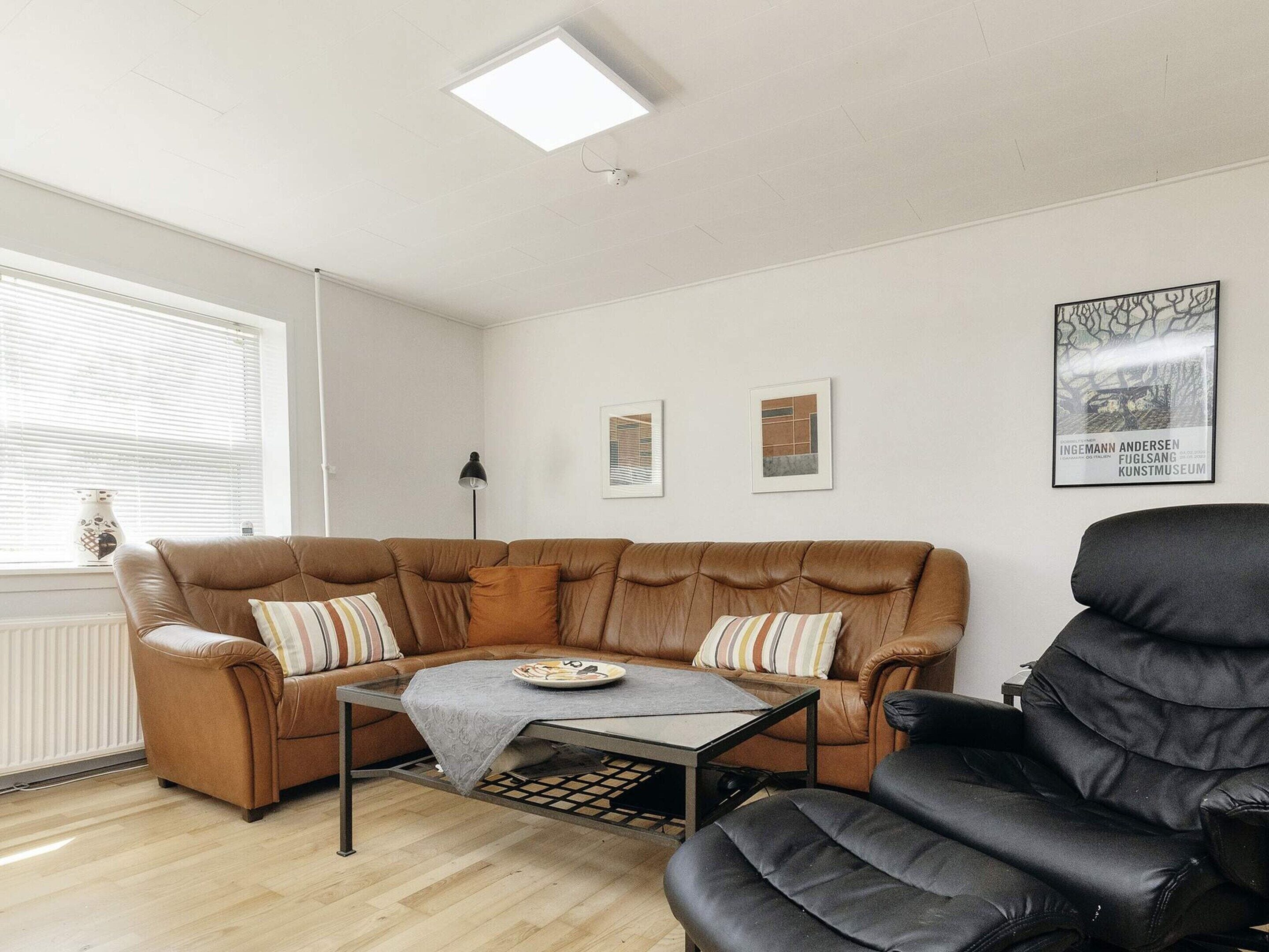A living room with a brown leather sofa, a black armchair, a glass coffee table, and a radiator. There are three framed artworks on the wall and a window with blinds.