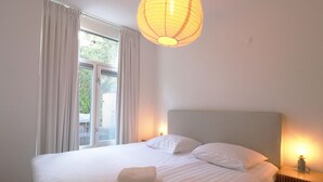 Een keurig opgemaakt bed met witte lakens en een opgevouwen handdoek, een nachtkastje met een lamp, en een raam met gordijnen.