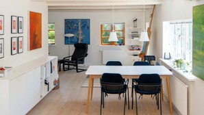 A dining area with a white table and four chairs, a black chair, a radiator, and a window with a view of greenery.