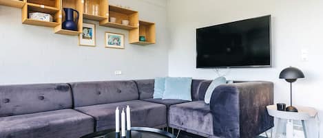 A modern living room with a sectional sofa, a flat-screen TV, wooden shelves, and a small side table.