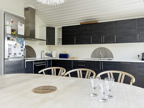 A modern kitchen with dark cabinets, a stainless steel refrigerator, a microwave, and a sink. There is a dining table with chairs and three wine glasses.